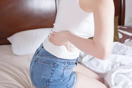 Woman In White Tank Top And Denim Shorts Sitting On Bed With Back Pain